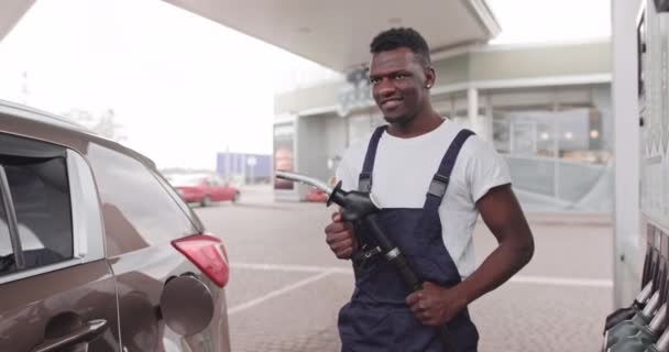 Jeune homme africain en vêtements de travail, travailleur de la station-service, est prêt à ravitailler la voiture, tenant la buse de pistolet de remplissage, regardant la caméra avec le sourire — Video