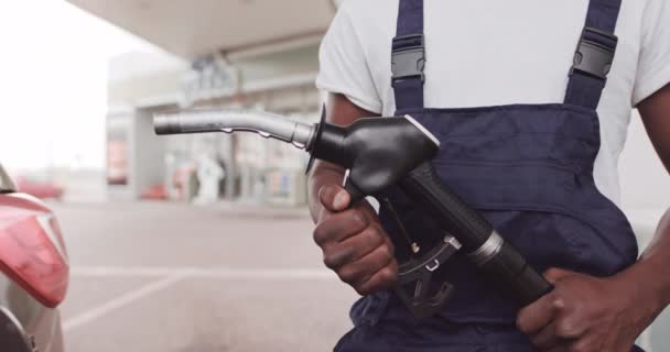 Imagen de cerca recortada del hombre de piel negra en ropa de trabajo, trabajador de la gasolinera, parado al aire libre en la gasolinera y sosteniendo la boquilla de la pistola de combustible en las manos — Vídeo de stock
