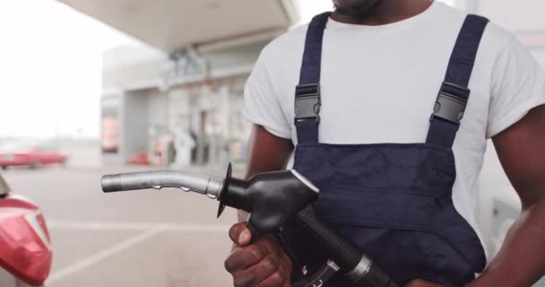 Bonito jovem africano, funcionário de posto de gasolina, usando t-shirt branca e macacão azul e segurando a arma de enchimento. Fechar vista frontal — Vídeo de Stock