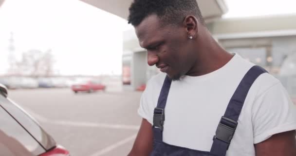 Petrol station, transportation and refueling concept. Handsome young black man, gas station worker, wearing dark blue overalls, refueling a car — Stock Video