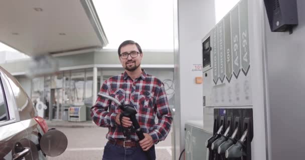 Jonge blanke man draagt casual outfit, is klaar om zijn luxe auto bij te tanken, houdt tankpistool mondstuk bij het tankstation, kijkt naar de camera met een glimlach. — Stockvideo