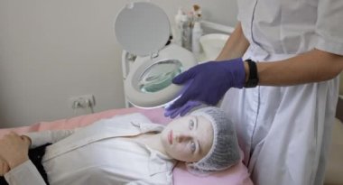 Kozmetik Kabinesi 'nde lambanın altında genç kadınların yakın çekimi. Yüz temizleme. Deri yenilenmesi ve su kaybı. Konsept güzellik merkezi, tıbbi klinik. Büyüteçle bak..