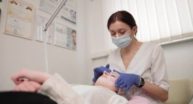 Koruyucu maskeli ve mavi lastik eldivenli genç bayan kozmetik uzmanı genç bayan müşteriler için yüz masajı ve cilt tedavisi yapıyor. Güzellik salonunda yüz masajı, yüz bakımı.
