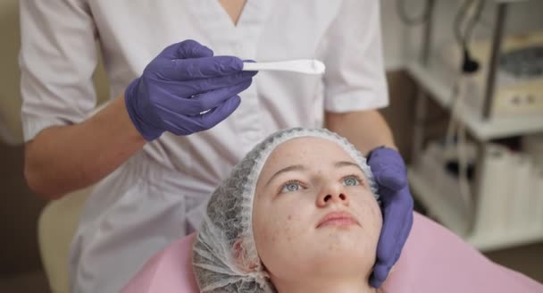 Нанесение пилинга на лицо. Молодая девушка в косметологическом кабинете проходит процедуру омоложения кожи лица. Косметология. Девушка косметолог работает с пациентами в офисе — стоковое видео