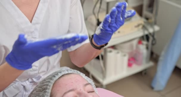 Cosmetologist mãos em luvas azuis tocando o rosto do paciente adolescente caucasiano fazendo procedimento de limpeza descascamento no salão de beleza. Acne, tratamento da pele, problema de pele. Conceito de cuidados com a pele. Lavagem facial — Vídeo de Stock