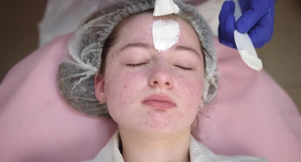 Cosmetologist händer tillämpa vit rengöring peeling lera mask på klientens ansikte med kosmetisk borste. Närbild av ung flicka i dermatologi klinik med hudbehandling förfarande — Stockvideo