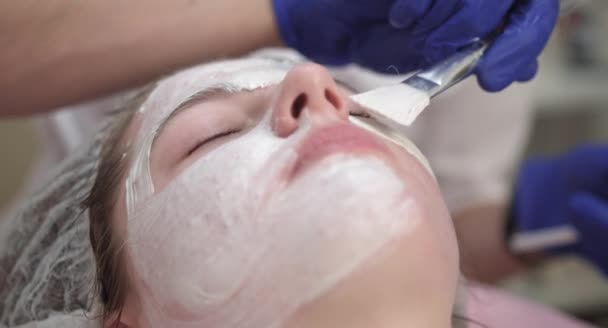 Cosmetologist hands applying white cleansing peeling mud mask on client face with cosmetic brush. Close up of young girl in dermatology clinic having skin treatment procedure — Stock Video