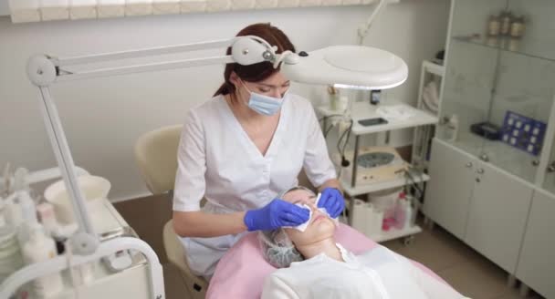 女性专业美容师的近视在蓝色手套清洁脸的少女患者用棉垫治疗痤疮。被胶卷覆盖的少女的脸。皮肤问题 — 图库视频影像