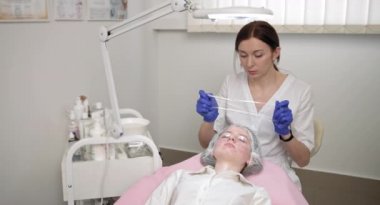 Kadın güzellik uzmanı müşterinin yüzüne lambadan bakıyor. Profesyonel kozmetoloji kliniğindeki genç kıza yaklaş. Sivilce tedavisi için plastik film ve maskeyle kaplı yüz..