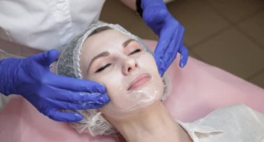 Kendine güvenen genç bayan kozmetik uzmanı, beyaz üniformalı ve mavi steril eldivenli, genç bayanın yüzüne temizleyici köpük sürüp, kanepede uzanan.