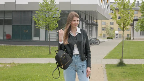 Joven estudiante, adolescente, en una chaqueta de cuero con una mochila, elegantemente vestida, corre mirando a la cámara y sonriendo. Movimiento muy lento. Retrato de mujer, caminar al aire libre en el parque, generación z — Foto de Stock