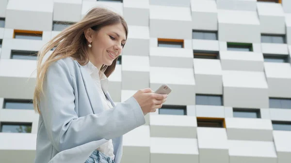 En forretningskvinne som bruker smarttelefon og sender e-post til en kvinnelig sjef. Forretninger. Sosialnettverk. Apps. Karrierefolk. Skyting på gaten nær kontoret. Jenta ser inn i kameraet og smiler. – stockfoto