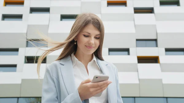Business nainen käyttää älypuhelin tekstailu naisten Executive tarkkailun sähköpostit. Työasioita. Sosiaalinen verkosto. Sovelluksia. Uraihmisiä. Ampumassa kadulla lähellä toimistoa. Tyttö katsoo kameraan ja hymyilee. — kuvapankkivalokuva