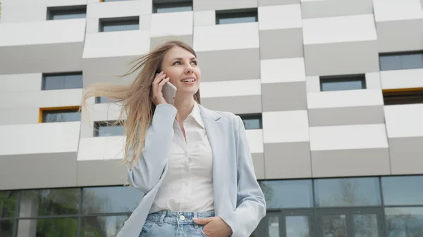 Hvit, selvsikker ung forretningskvinne i hvit skjorte snakker i telefonen utenfor moderne kontorbygg. Medium lang-lav-vinkel 4K sakte bevegelses korporat skutt med beveger seg rundt. Karriere – stockfoto