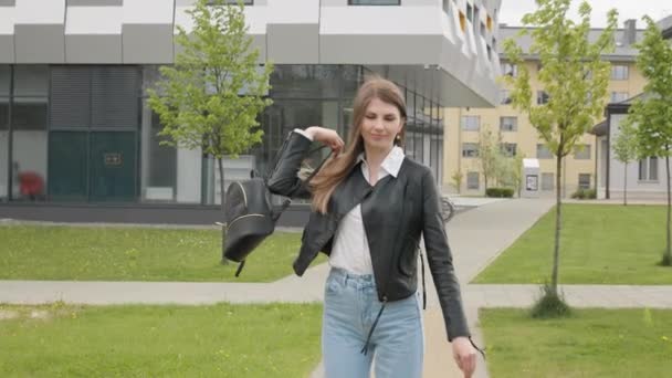 Jovem estudante, adolescente, em uma jaqueta de couro com uma mochila, elegantemente vestida, corre olhando para a câmera e sorrindo. Movimento muito lento. Mulher retrato, caminhando ao ar livre no parque, geração z — Vídeo de Stock