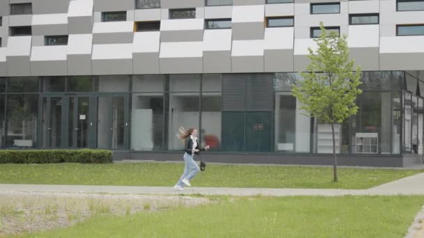 Молодая девушка студентка, подросток, в кожаной куртке с рюкзаком, стильно одетая, бежит рядом с современным офисом или зданием колледжа. Очень медленное движение, улыбка в камеру. Счастливая девочка-подросток — стоковое видео