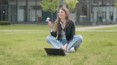 Genç, şık giyimli bir kız bilgisayarın yanındaki çimlerin üzerinde oturmuş elinde bir karahindiba tutuyor ve çiçekleri üflüyor. Ağır çekim. Şehir parkında çimlerde otururken dizüstü bilgisayarda çalışan meşgul bir kız.