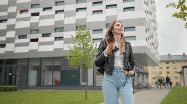 Şık giyimli bir kız, kotlar ve deri ceketle dışarıda eğleniyor. Mutlu ve hoş görünüyor. Şık görünüyorsun. Binanın arka planında bir kız öğrencinin portresi var. Güzel bir makyajla