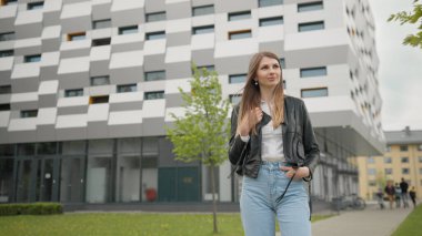 Şık giyimli bir kız, kotlar ve deri ceketle dışarıda eğleniyor. Mutlu ve hoş görünüyor. Şık görünüyorsun. Binanın arka planında bir kız öğrencinin portresi var. Güzel bir makyajla