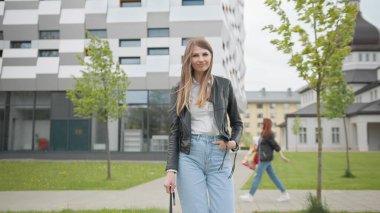 Sırt çantalı kız üniversite binasına doğru yürüyor. Mutlu ve gülümsüyor, kameraya bakıyor ve ileri doğru yürüyor. Beyaz tişörtlü Bayan Öğrencinin Portresi Kameraya Emin Bir şekilde Bakıyor