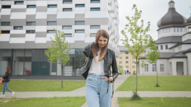 Sırt çantalı kız üniversite binasına doğru yürüyor. Mutlu ve gülümsüyor, kameraya bakıyor ve ileri doğru yürüyor. Beyaz tişörtlü Bayan Öğrencinin Portresi Kameraya Emin Bir şekilde Bakıyor