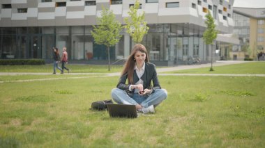 Genç bir kadın, kulaklıkla müzik dinliyor, akıllı telefon kullanıyor, gün batımında yürüyor, gülümsüyor, yavaş çekimde etrafına bakıyor. Okulun, üniversitenin ya da ofisin yakınındaki çimlerde oturan çekici kız öğrenci.