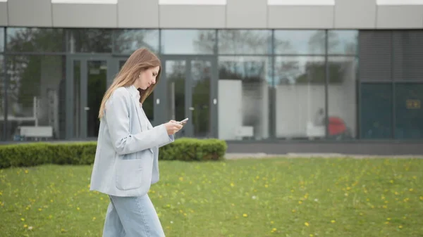 En glad ung dame som får rabatt for å handle mens hun sjekker e-postkassen mens hun går nedover gaten. Kvinne taster inn kredittkortnummer til nettshopping med smarttelefon – stockfoto