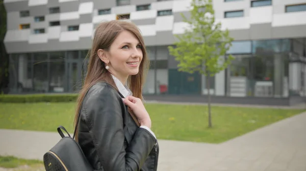 Um retrato de uma jovem, um estudante perto de uma faculdade ou escritório moderno, andando pela rua com uma mochila. Elegantemente Vestida Moderna Menina Lentamente Coloca um saco de ombro. Tiroteio pelas costas — Fotografia de Stock