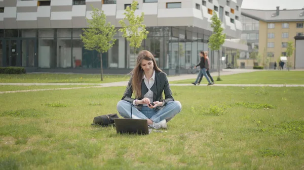 Teini nainen, musiikin kuuntelu kuulokkeissa käyttää älypuhelinta kaupungin kävelyllä auringonlasku katsoa ympärilleen hymy muotokuva, hidastettuna. Houkutteleva naispuolinen opiskelija istuu nurmikolla koulun tai yliopiston tai toimiston lähellä — kuvapankkivalokuva