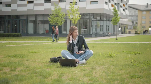 ผู้หญิงวัยรุ่น ฟังเพลงในหูฟัง ใช้สมาร์ทโฟนตอนพระอาทิตย์ตก มองไปรอบๆ ภาพยิ้ม การเคลื่อนไหวช้า นักเรียนหญิงที่น่าสนใจนั่งบนหญ้าใกล้โรงเรียนหรือวิทยาลัยหรือออฟฟิศ — ภาพถ่ายสต็อก