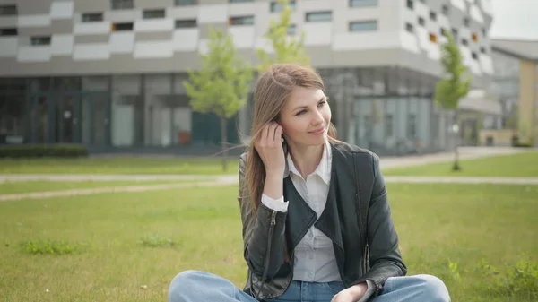 Houkutteleva naispuolinen opiskelija istuu nurmikolla koulun tai yliopiston tai toimiston lähellä. Teini nainen, musiikin kuuntelu kuulokkeissa käyttää älypuhelinta kaupungin kävelyllä auringonlasku katso ympärillesi hymy muotokuva, hidastettuna — kuvapankkivalokuva
