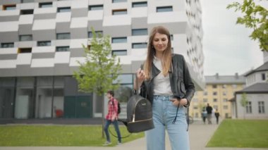 Şık giyimli bir kız, kotlar ve deri ceketle dışarıda eğleniyor. Mutlu ve hoş görünüyor. Şık görünüyorsun. Binanın arka planında bir kız öğrencinin portresi var. Güzel bir makyajla