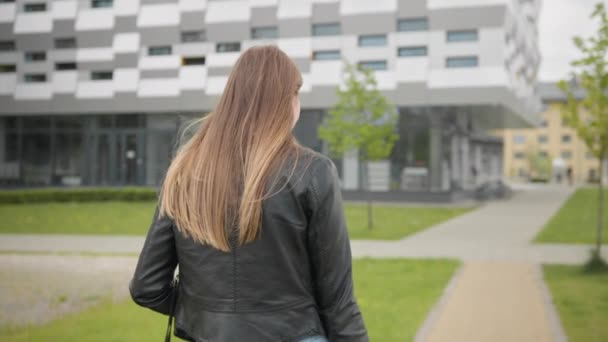 Um retrato de uma jovem, um estudante perto de uma faculdade ou escritório moderno, andando pela rua com uma mochila. Elegantemente Vestida Moderna Menina Lentamente Coloca um saco de ombro. Tiroteio pelas costas — Vídeo de Stock