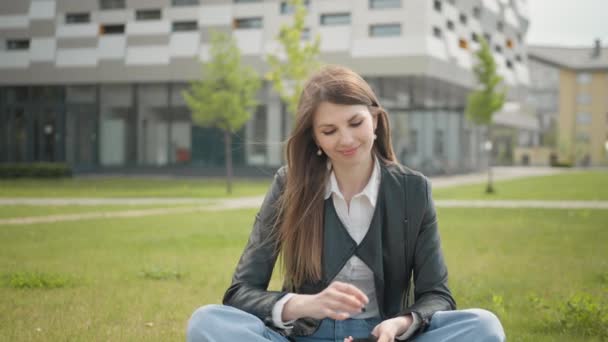 Ελκυστική φοιτήτρια που κάθεται στο γρασίδι κοντά στο σχολείο ή στο κολέγιο ή στο γραφείο. Έφηβη γυναίκα, ακούγοντας μουσική σε ακουστικά χρήση smartphone στο ηλιοβασίλεμα βόλτα στην πόλη ματιά γύρω από το χαμόγελο πορτρέτο, αργή κίνηση — Αρχείο Βίντεο
