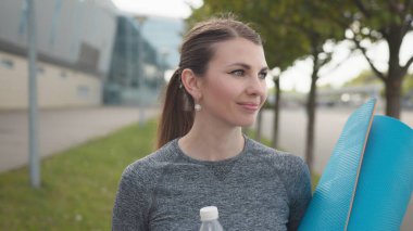 Neşeli, hoş bir genç kadının portresi, yaz mevsimi pozitif esmer kız şehir manzarasında spor yaptıktan sonra dinleniyor. Ellerinde halısı ve suyu olan kız, Kameraya Yavaşça Bak ve Gülümse.