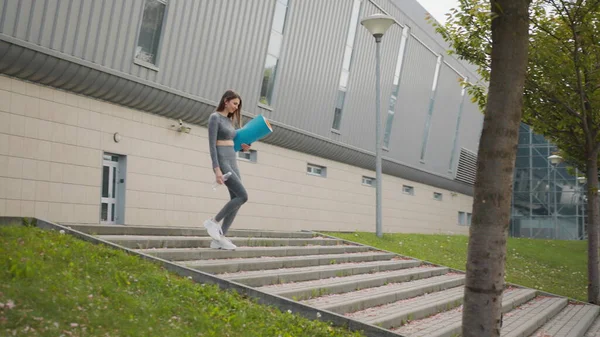 Ung sportig vacker flicka med en matta i händerna och vatten, kör uppför trappan i centrum. Vacker tonåring går till träningen. Begreppet idrott, hälsosam livsstil, viktminskning. — Stockfoto
