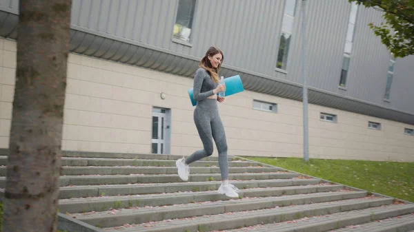 Flickan springer nerför trappan i slow motion. Löpare kvinna som springer på trappor vid utomhus träning. Ung sportig vacker flicka med en matta i händerna och vatten, kör uppför trappan i centrum — Stockfoto