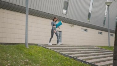 Elinde halı ve suyla şehir merkezindeki merdivenlerden yukarı koşan genç ve güzel bir kız. Güzel Genç Kız Antrenmana Gidiyor. Spor anlayışı, sağlıklı yaşam tarzı, kilo kaybı.