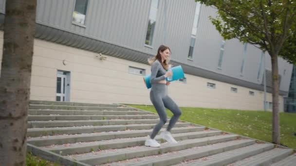 Flickan springer nerför trappan i slow motion. Löpare kvinna som springer på trappor vid utomhus träning. Ung sportig vacker flicka med en matta i händerna och vatten, kör uppför trappan i centrum — Stockvideo