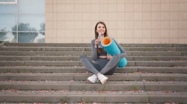 Eğitimden ve içme suyundan sonra merdivenlerde oturan genç ve güzel atletik kız. İyi ruh hali ve sağlıklı yaşam konsepti. Fitness anlayışı, sağlıklı yaşam tarzı. Antrenmandan sonra merdivenlerde oturan kadın.