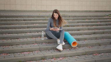 Fitness Kadını telefonda egzersiz uygulaması kullanıyor. Merdivenlerde oturarak egzersiz yapıyor. Spor ve Teknoloji, Kilo Verme ve Eğitim. Spor salonunda dinlenirken cep telefonu kullanan kadın