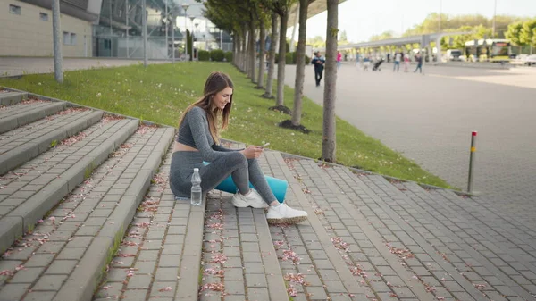 Fitness nainen käyttää puhelinta Workout App liikunta istuu kaupungin portaissa. Urheilu ja teknologia, laihtuminen ja koulutus. Nainen, joka käyttää matkapuhelinta levätessään kuntoilun aikana — kuvapankkivalokuva
