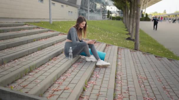 Mujer Fitness Usando Teléfono Con Aplicación de Entrenamiento Ejercicio Sentado en la Ciudad en las escaleras. Deporte y tecnología, pérdida de peso y entrenamiento. Mujer usando teléfono móvil mientras descansa durante el entrenamiento de fitness — Vídeos de Stock