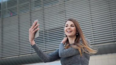 Kafkasyalı genç kadının modern binanın arka planında görüntülü konuşma yaparken yakın plan. Açık havada. Çekici ve mutlu kızın POV 'u. Akıllı Telefonu Tutan Güzel Bir Spor Kızının Yakın Çekimi