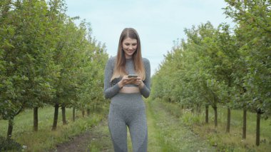 Elinde telefon tutan genç bir atletik fitness kızının portresi. Sokakta ağaçların arasındaki piraturada ateş etmek. Kız telefondan bir mesaj okuyor ve programı kullanıyor, gülümsüyor.