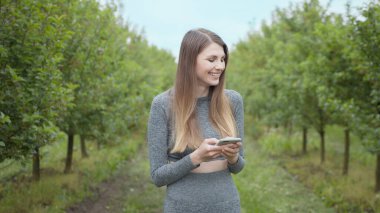 Dışarıda cep telefonuyla yazan mutlu kadın. Arka planda akıllı telefonu olan neşeli bir kız. Dışarıda cep telefonunu tutan gülümseyen kadın. Atletik fitness kızı elinde telefon tutuyor..