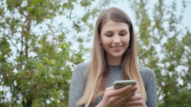Dışarıda cep telefonuyla yazan mutlu kadın. Arka planda akıllı telefonu olan neşeli bir kız. Dışarıda cep telefonunu tutan gülümseyen kadın. Spor kızı, internet teknolojisi, koşucu egzersizi.