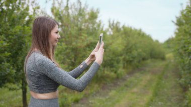 Akıllı telefondaki fotoğraf, arka plandaki ağaçlar, güzel bir kızın akıllı telefondaki bahar ağaçlarını fotoğraflaması. Ortalama bir plan. Dışarıda cep telefonuyla yazan mutlu kadın. Parktaki neşeli kız.