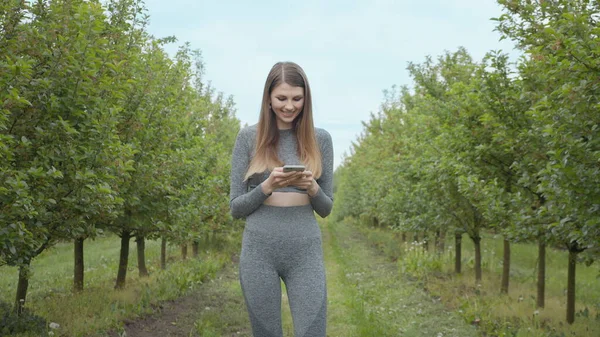 Ritratto di una giovane atleta che tiene un telefono tra le mani. Sparatoria sulla strada sul pirodo tra gli alberi. La ragazza legge un messaggio dal telefono e usa il programma, sorridendo — Foto Stock