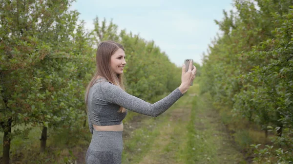 Ung attraktiv sport flicka skytte på ett telefonträd fotograferad mot naturen bakgrund. Flicka bloggare pekar på kameran naturen, begreppet berättelser om miljön skydd av träd — Stockfoto